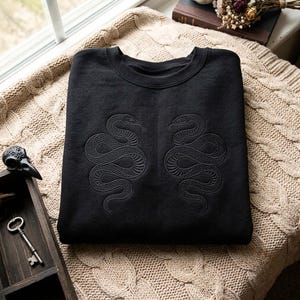 May include: A black crewneck sweatshirt featuring two symmetrical, tone-on-tone embroidered snakes facing each other on the chest. The snakes are depicted in a coiled position with visible scale detailing, creating a subtle, textured design on the fabric.