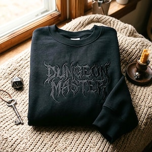Puede incluir: Una sudadera negra de cuello redondo con las palabras Dungeon Master bordadas en el pecho en una fuente oscura, estilizada e inspirada en el estilo gótico. La prenda tiene mangas largas con puños acanalados y un cuello acanalado en negro.