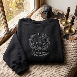 Pode incluir: Uma camisola preta de gola redonda com bordado branco de três corvos pousados num ramo dentro de uma coroa circular de folhas, estrelas e cristais. Abaixo do desenho, o texto diz "SUPPORT YOUR LOCAL MURDER" numa fonte simples.