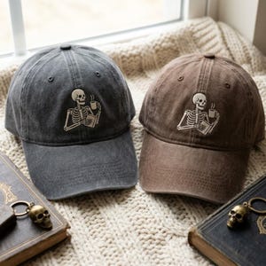 May include: Two baseball caps, one charcoal gray and one brown, each featuring an embroidered graphic of a skeleton holding a steaming mug and a book. The caps have a distressed, washed finish, a curved brim, and a button on the top crown.