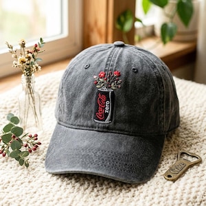 May include: A charcoal gray baseball cap featuring an embroidered design of a black soda can labeled Coca-Cola zero with red flowers emerging from the top. The cap has a structured crown, a curved brim, and visible stitching details throughout the fabric.