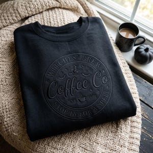 May include: A black crewneck sweatshirt featuring a tonal embossed circular graphic that reads Witches' Brew Coffee Co, Est. 1692, and Stop In For A Spell, with illustrations of a witch on a broomstick, bats, stars, and a coffee cup.