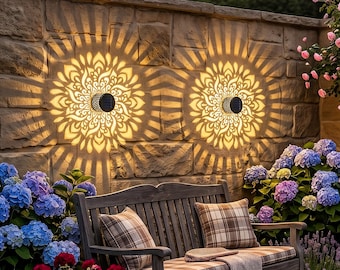 May include: Two solar-powered wall lights with intricate floral patterns cast a warm glow on a stone wall. A wooden bench with plaid cushions sits below, surrounded by colorful flowers, creating a serene outdoor scene.
