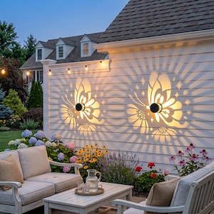 May include: Outdoor wall lights casting butterfly-shaped light patterns. The lights are mounted on a white wall, creating a decorative effect. The scene includes outdoor seating, plants, and string lights, suggesting a garden setting.