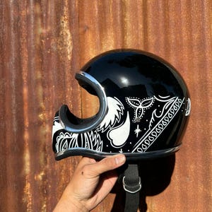 May include: A glossy black motorcycle helmet featuring a white hand-painted paisley pattern across the side. The helmet has a chrome-coloured trim around the face opening and a black chin strap with a metal D-ring closure system.