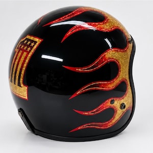 Può includere: Un casco da moto nero lucido con un motivo grafico a fiamme dorate e rosse sul lato. Il casco presenta un prominente emblema dorato a forma di numero uno con piccole stelle integrate nella parte superiore della cifra sul lato.