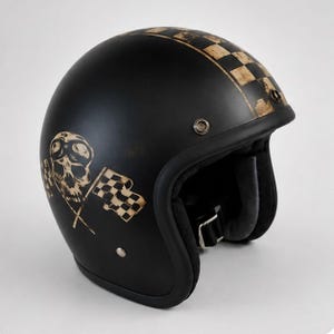 May include: A matte black open-face motorcycle helmet featuring a distressed gold graphic of a skull wearing goggles crossed with two chequered flags on the side and a matching chequered racing stripe running over the top of the helmet shell.