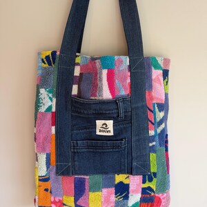 May include: A tote bag constructed from a patchwork of colourful, textured terry cloth fabric squares. The bag features dark blue denim straps and a central dark blue denim pocket panel with a white label displaying the text ZOLVI and a wave icon.