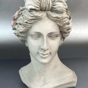 May include: A gray stone-colored bust of a person with a serene expression, featuring wavy hair styled with a prominent bow on top and small, muted pink and green floral accents tucked into the sides of the hair, set against a neutral background.