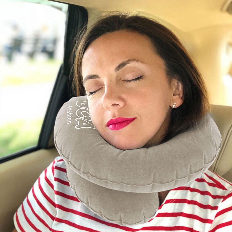 May include: A gray inflatable travel pillow with the text "BCA cushion" is around a person's neck. The person is wearing a red and white striped shirt and has closed eyes. The background is blurred, suggesting a car interior.