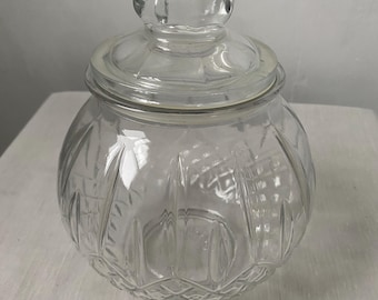 May include: A clear glass jar with a matching lid featuring a rounded shape and a textured pattern. The body of the jar displays vertical panels and a diamond-cut design at the base, while the lid includes a knob handle for easy removal.
