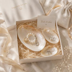 May include: A gift box containing three iridescent shell-shaped dishes nestled in shredded paper. The dishes hold a pearl bracelet, two silver rings, and a small silver charm. A card reads "Best Wishes" with a heart, and the box lid says "Especially for you".