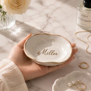 May include: A small, round, off-white ceramic trinket dish with an irregular, wavy rim and a speckled finish. The center of the dish features the name Miller printed in a dark brown, elegant cursive script font on the smooth interior surface.