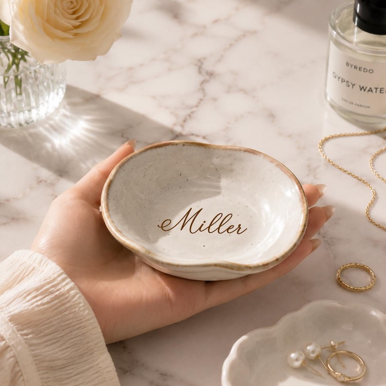 May include: A small, round, off-white ceramic trinket dish with an irregular, wavy rim and a speckled finish. The center of the dish features the name Miller printed in a dark brown, elegant cursive script font on the smooth interior surface.