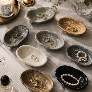 May include: A collection of eight small, oval-shaped ceramic trinket dishes in various colors including white, grey, black, and tan. Each dish features a textured, organic rim and holds various pieces of gold jewelry and pearl bracelets.