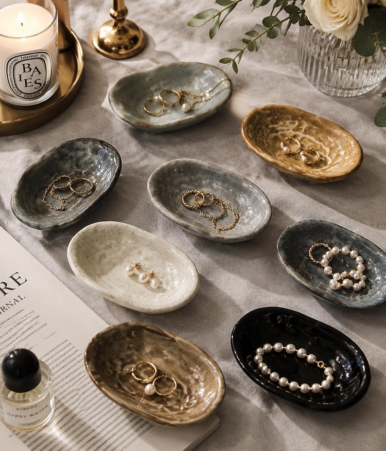 May include: A collection of eight small, oval-shaped ceramic trinket dishes in various colors including white, grey, black, and tan. Each dish features a textured, organic rim and holds various pieces of gold jewelry and pearl bracelets.
