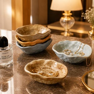 May include: A collection of small, irregular, organic-shaped ceramic bowls in muted earth tones including beige, ochre, and light blue. Some bowls are stacked, while others hold gold jewelry pieces like necklaces, rings, and a watch.