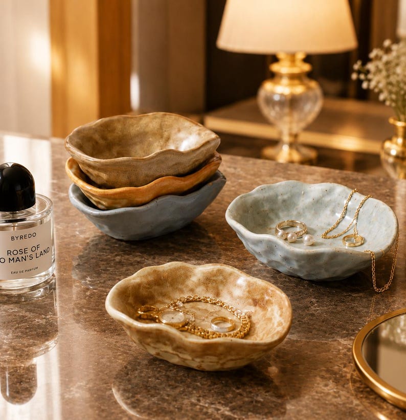 May include: A collection of small, irregular, organic-shaped ceramic bowls in muted earth tones including beige, ochre, and light blue. Some bowls are stacked, while others hold gold jewelry pieces like necklaces, rings, and a watch.