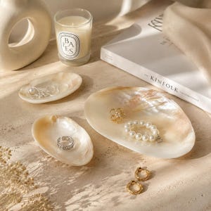 May include: Three iridescent, oval-shaped shell dishes used as jewelry trays. The dishes hold a silver chain, a silver ring, a gold flower-shaped brooch, and a white pearl bracelet. Two gold hoop earrings lie nearby on the surface.