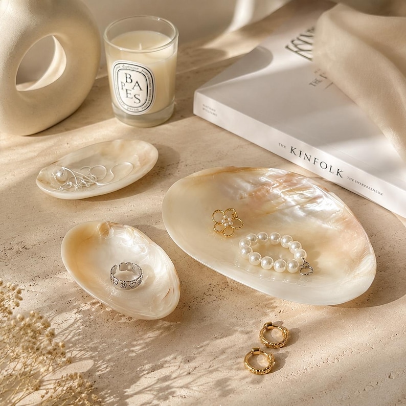 May include: Three iridescent, oval-shaped shell dishes used as jewelry trays. The dishes hold a silver chain, a silver ring, a gold flower-shaped brooch, and a white pearl bracelet. Two gold hoop earrings lie nearby on the surface.