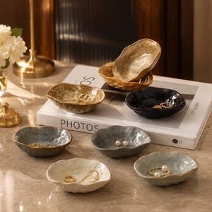 May include: A collection of seven small, irregular, organic-shaped ceramic bowls in various colors including gold, black, gray, and off-white. These bowls feature textured, speckled surfaces and uneven, wavy rims, some containing small pieces of jewelry.