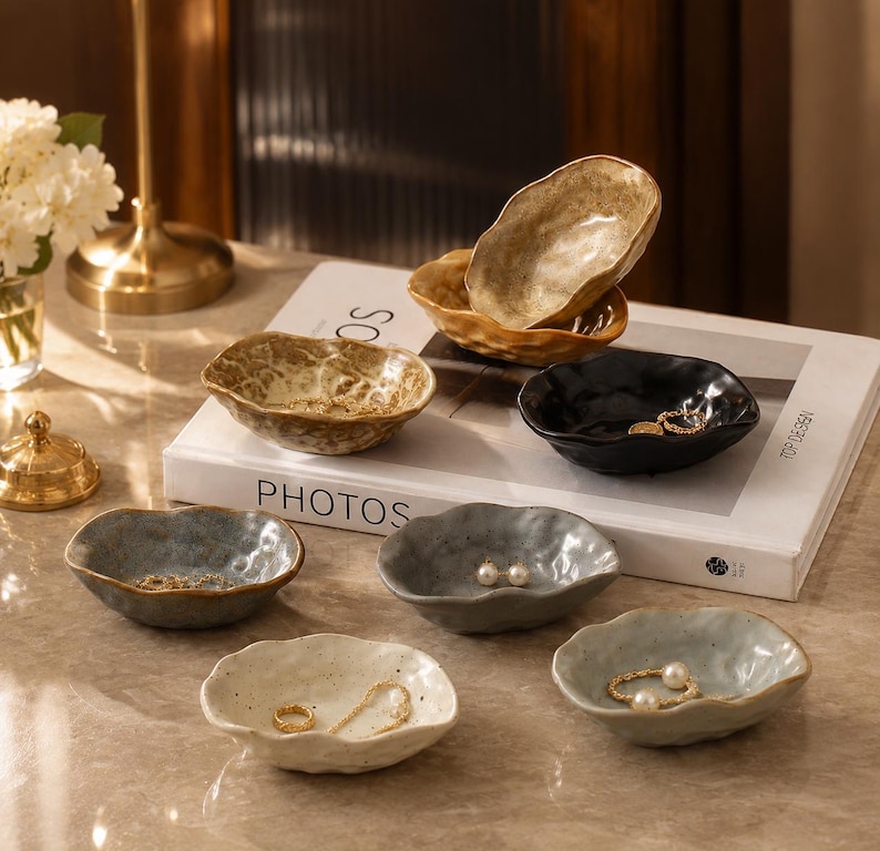 May include: A collection of seven small, irregular, organic-shaped ceramic bowls in various colors including gold, black, gray, and off-white. These bowls feature textured, speckled surfaces and uneven, wavy rims, some containing small pieces of jewelry.