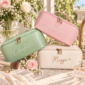 May include: Three rectangular cosmetic bags with top handles and gold-tone double zippers. One bag is pastel pink with the name Amelia, one is sage green with the name Charlotte, and one is cream with the name Maggie and a small heart icon.