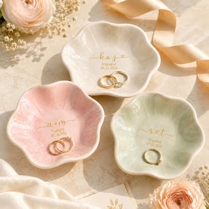 May include: Three flower-shaped ceramic ring dishes in white, soft pink, and sage green. Each dish features gold-colored text with initials, the word Engaged, and a date. The white dish reads k & j 06.21.2026, the pink a + m 05.18.2026, and the green s & t 07.12.2026.