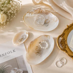 May include: Three iridescent, oval-shaped shell dishes used as jewelry trays. The largest dish holds a beaded bracelet and a ring. A smaller dish contains a delicate earring. The third dish holds a pearl bracelet and a pair of gold-toned earrings.