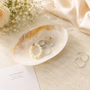 May include: An iridescent shell-shaped trinket dish containing a pearl bracelet, a gold-toned flower ring, and a silver chain. Beside the dish are two silver hoop earrings and a white card with the text YESTERDAY, today is a gift, tomorrow is mystery.