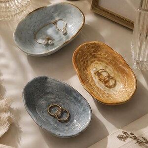 May include: Three small, oval-shaped ceramic trinket dishes with irregular, organic edges. One dish is light blue with dark speckles, one is a deeper grey-blue, and one is a warm, mottled tan. Each dish holds gold-toned jewelry pieces.