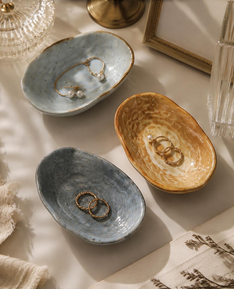 May include: Three small, oval-shaped ceramic trinket dishes with irregular, organic edges. One dish is light blue with dark speckles, one is a deeper grey-blue, and one is a warm, mottled tan. Each dish holds gold-toned jewelry pieces.