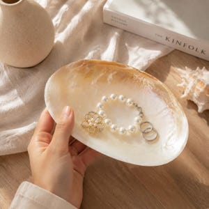 May include: An oval-shaped, iridescent mother-of-pearl shell dish containing a white pearl bracelet, a gold-toned flower-shaped brooch with small white pearls, and two thin silver-toned metal rings resting inside the smooth, concave surface.