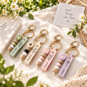 Op de afbeelding: Vier gepersonaliseerde sleutelhangers met goudkleurige metalen ringen en sluitingen. Elke hanger heeft een platte, leerachtige band met een florale lijntekening en een naam: Jennifer, Analise, Diane of Talya, inclusief een bloemvormige kraal.