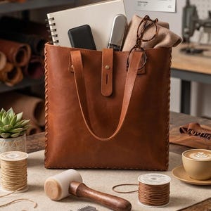 May include: A brown leather tote bag with hand-laced edges, twin straps, and a center tuck-tab closure. The bag contains a spiral notebook, a black case, a silver tool, and tortoiseshell glasses. A paper sign reads: a hand-laced leather tote.