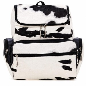Puede incluir: Una mochila con estampado de manchas de vaca en blanco y negro y un acabado texturizado de pelo. La bolsa incluye un compartimento principal grande con solapa superior, un bolsillo frontal con cremallera y detalles en negro con cremalleras de metal plateado.