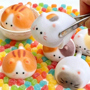 May include: A collection of round, squishy cat-shaped toys in various colors including orange tabby, white with calico patches, and gray. One white cat toy is being held by metal tongs. The toys feature small ears, whiskers, and black eyes.