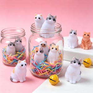 May include: A collection of small, flocked cat figurines in various colors including white, gray, and orange, displayed inside and around two clear glass jars filled with colorful shredded paper, accompanied by two small yellow metal bells.