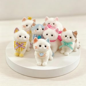 May include: A collection of seven small, white, flocked cat figurines. Each cat features unique accessories, including a gold crown, a yellow gingham bib, a blue gingham collar, a pink ribbon, and a teal bow. One cat wears a heart-shaped pendant.