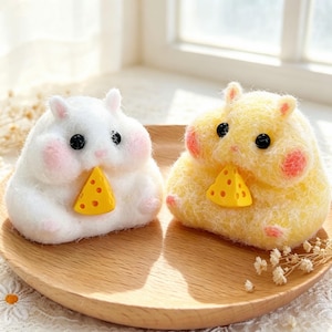 May include: Two needle-felted hamster figurines, one white and one yellow, each featuring small black bead eyes, pink cheeks, and holding a small yellow triangular piece of cheese with holes. The figures have a fuzzy texture and rounded shapes.