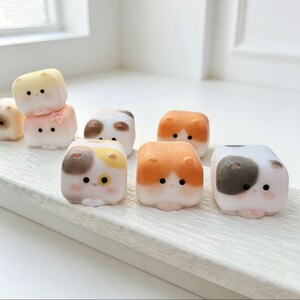 May include: A collection of small, cube-shaped figurines resembling cats with rounded ears and simple facial features. The figures are rendered in various colours including white, orange, brown, and yellow, with some featuring small pink bows or spots.