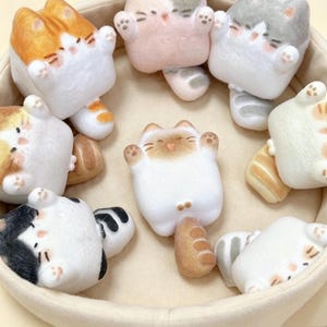 May include: A collection of small, plush, cat-shaped figures with rounded bodies and raised paws. The figures feature various color patterns including orange, grey, black, and white, with simple facial expressions and tails, arranged inside a circular container.