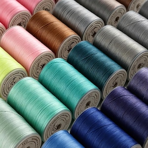 Peut inclure: Gros plan de plusieurs bobines de fil de différentes couleurs, dont rose, marron, sarcelle et bleu. Les bobines sont disposées en rangées soignées, présentant la variété de couleurs disponibles pour les projets de couture et d'artisanat.