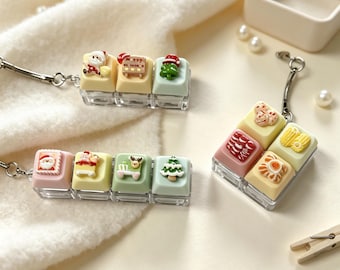 May include: Three keychains featuring miniature pastel-colored mechanical keyboard keycaps decorated with Christmas-themed charms. The charms include Santa Claus, a sleigh, a reindeer, a Christmas tree, and various holiday cookies in red, green, and yellow.