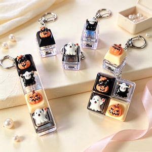 May include: A collection of Halloween-themed keycap keychains featuring miniature figures of orange jack-o'-lanterns, white ghosts, and black figures with white hats. The keycaps are mounted on clear bases with silver metal key rings attached.