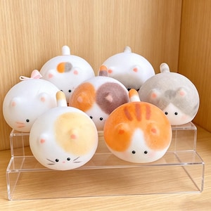 May include: A collection of seven round, cat-shaped figurines with smooth, matte surfaces. Each figurine features small ears, tiny black eyes, and distinct color patterns including orange, gray, and calico patches, displayed on a clear tiered stand.