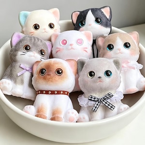 May include: A collection of seven small, flocked cat figurines with large, glossy eyes, arranged inside a white bowl. The cats feature various colors including white, gray, brown, and black, with some wearing small bows, collars, or ruffled collars.