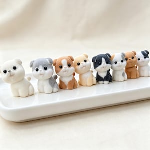 May include: A collection of eight small, flocked animal figurines arranged in a row on a white rectangular tray. The figures include various puppy and kitten designs in shades of white, gray, tan, and black, each featuring large black eyes.