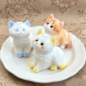 May include: Three small cat figurines with fuzzy textures and tiny silver halos on their heads. One cat is white with blue ears and paws, one is white with yellow ears and paws, and one is orange and white tabby, all featuring dark eyes.