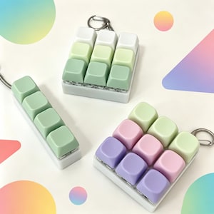以下が含まれることがあります： メカニカルキーボードのキーキャップを使用した3つのキーホルダー。1つは白い長方形のベースに4つのセージグリーンのキー。もう1つは白い正方形のベースに白とセージグリーンの9つのキー。3つ目は白い正方形のベースにパステルカラーの9つのキーが付いています。